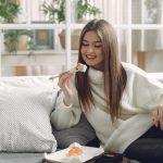 Smiling young lady in warm knitted sweater enjoying fresh sushi on wicker tray while relaxing on cozy sofa with soft cushions in modern living room