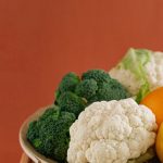 Close-up of fresh broccoli, cauliflower, lemon, and orange on a wooden plate with an earthy background.